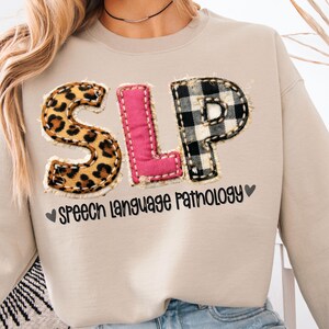 May include: Beige sweatshirt with the letters "SLP" in leopard print, pink, and black and white buffalo plaid. The text "Speech Language Pathology" is below the letters, with a heart symbol on each side.