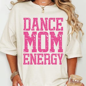 May include: Cream-colored t-shirt with the words "DANCE MOM ENERGY" in distressed pink lettering. The shirt has a relaxed fit and short sleeves. A woman is wearing the shirt and holding a glass of iced coffee.