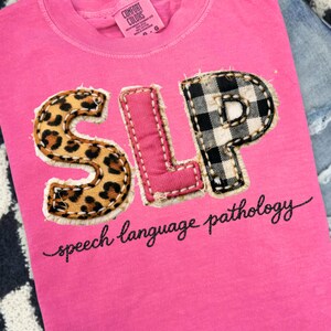 May include: A bright pink t-shirt with the letters "SLP" appliqued on the front. The "S" has a leopard print, the "L" is pink with white stitching, and the "P" is black and white buffalo plaid. The words "speech language pathology" are written below.