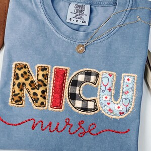 Puede incluir: Una camiseta azul claro con las letras "NICU" en un diseño de patchwork. La "N" es de estampado de leopardo, la "I" es roja, la "C" es a cuadros blancos y negros, y la "U" tiene un tema médico. La palabra "nurse" está bordada en rojo.