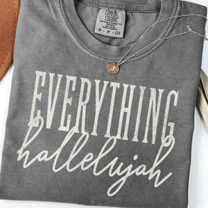 Op de afbeelding: Een grijs Comfort Colors T-shirt met de woorden "EVERYTHING hallelujah" in witte letters. Een zilveren ketting met een kleine hanger ligt op het shirt. Het shirt is netjes opgevouwen.
