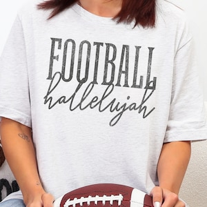 Op de afbeelding: Een lichtgrijs gem&ecirc;leerd t-shirt met de tekst FOOTBALL hallelujah in een verweerd zwart lettertype. Het woord FOOTBALL is gedrukt in een vet, blokkerig schreefloos lettertype, terwijl het woord hallelujah in een cursief handschrift staat.