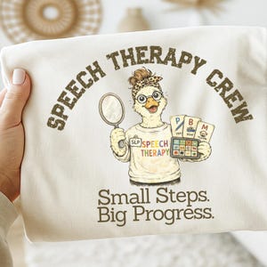 Könnte beinhalten: Cremefarbenes Sweatshirt mit dem Text "SPEECH THERAPY CREW" und "Small Steps. Big Progress." Eine Cartoon-Ente mit Brille und Stirnband hält einen Spiegel und Karten mit Sprachtherapie-Symbolen.