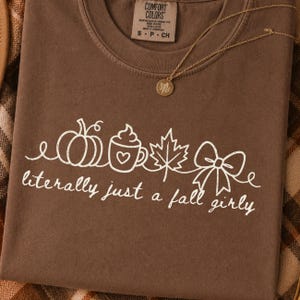 May include: A brown Comfort Colors t-shirt with a white graphic of a pumpkin, coffee cup, maple leaf, and bow. The text "literally just a fall girly" is below the graphic. A gold necklace with a leaf pendant is on the shirt.