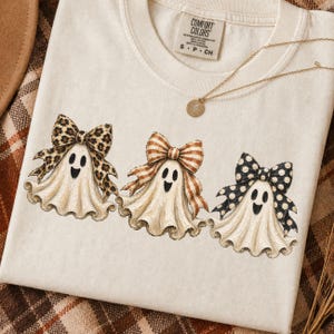 May include: Cream-colored t-shirt featuring three cartoon ghosts with patterned bows. The bows have leopard print, striped, and polka dot designs. A gold necklace with a pendant is also visible.