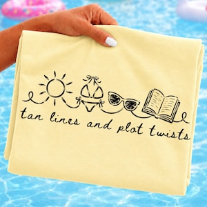 Op de afbeelding: Een gevouwen, lichtgele strandlaken met een zwarte afbeelding van een zon, bikini, zonnebril en een open boek. De tekst "tan lines and plot twists" staat eronder. De hand houdt de strandlaken vast.