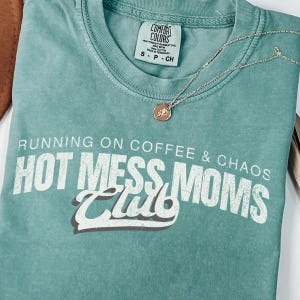 May include: A seafoam green t-shirt with the text "RUNNING ON COFFEE & CHAOS HOT MESS MOMS Club" in white. A gold-colored necklace with a small pendant rests on the shirt. The shirt has a Comfort Colors tag.