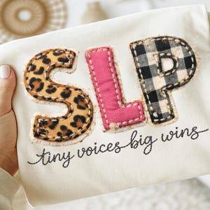 Könnte beinhalten: Cremefarbener Stoff mit den Buchstaben "SLP", appliziert in Leopardenmuster, Rosa und schwarz-weißem Büffelleder. Der Satz "tiny voices big wins" ist in schwarzer Schrift unter den Buchstaben gestickt.