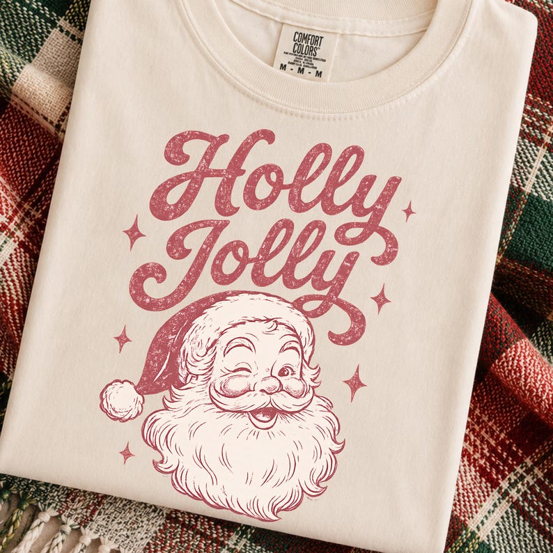 May include: A cream-colored t-shirt featuring a distressed red graphic of a winking Santa Claus and the text "Holly Jolly" in a cursive font. The shirt has a Comfort Colors brand label at the neck with a size M tag visible on the collar.