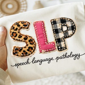 May include: Cream-colored fabric with the letters "SLP" appliqued in leopard print, pink, and black and white plaid. The words "speech language pathology" are written in cursive below the letters.