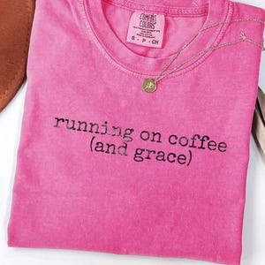 Puede incluir: Una camiseta rosa fucsia doblada con la frase "running on coffee (and grace)" impresa en negro. Un collar dorado con un pequeño colgante descansa sobre la camiseta. La camiseta tiene una etiqueta Comfort Colors.