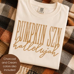 May include: A cream-colored short-sleeve t-shirt featuring the text PUMPKIN SZN in a distressed gold serif font, with the word hallelujah written below in a gold cursive script. The shirt has a Comfort Colors brand label at the neck.