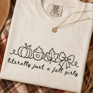 May include: Cream-colored t-shirt with a black line drawing of a pumpkin, coffee cup, maple leaf, and bow. The text "literally just a fall girly" is below the drawing. A gold necklace with a round pendant is on the shirt.