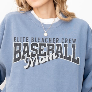 Puede incluir: Sudadera azul claro con el texto "ELITE BLEACHER CREW BASEBALL Mom" en un estilo vintage. La sudadera tiene cuello redondo y mangas largas. Se ve una camiseta blanca debajo.
