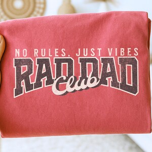 May include: A red t-shirt with the text "NO RULES, JUST VIBES" above the words "RAD DAD Club" in a distressed, vintage-style font. The shirt is folded, and held by a hand.