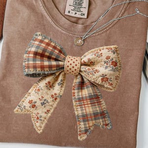 May include: A brown t-shirt with a large, decorative bow appliqué. The bow features a mix of patterns, including plaid, floral, and polka dots, in shades of red, beige, and blue. A silver necklace with a pendant is also visible.