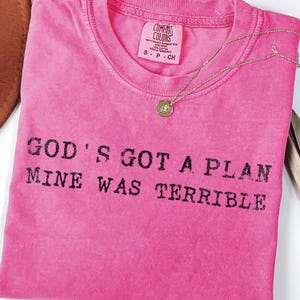 May include: A vibrant pink t-shirt featuring the black text "GOD'S GOT A PLAN MINE WAS TERRIBLE". The shirt has a crew neck and a small label. A gold necklace with a pendant is placed on the shirt.