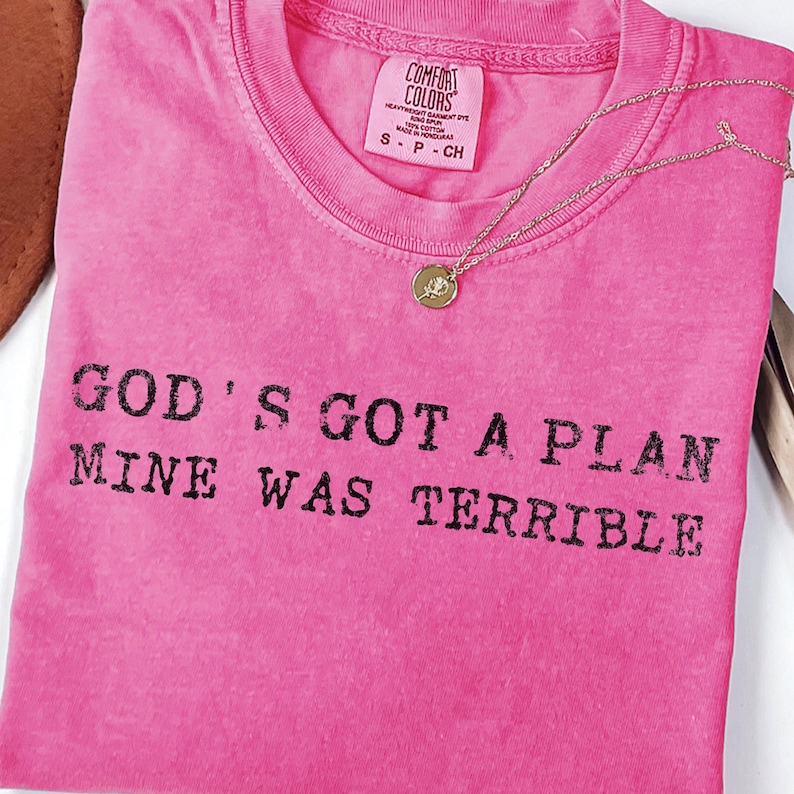 May include: A vibrant pink t-shirt featuring the black text "GOD'S GOT A PLAN MINE WAS TERRIBLE". The shirt has a crew neck and a small label. A gold necklace with a pendant is placed on the shirt.