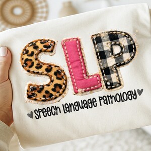 May include: Cream-colored fabric with the letters "SLP" in a patchwork design. The "S" is leopard print, the "L" is pink, and the "P" is black and white checkered. The words "Speech Language Pathology" are printed below.
