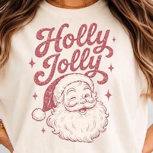 May include: A cream-colored t-shirt featuring a distressed red graphic of a winking Santa Claus wearing a hat. Above the Santa illustration, the text "Holly Jolly" is printed in a bold, cursive, distressed red font, surrounded by small stars.