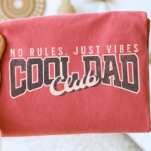 May include: A red t-shirt with the text "NO RULES, JUST VIBES COOL DAD Club" in a vintage, collegiate-style font. The text is in a combination of white, brown, and red colors. The shirt is folded.