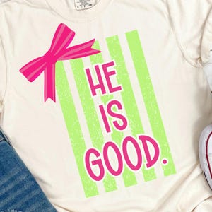 Pode incluir: Camiseta creme com laço rosa e design de listras verdes. As palavras "HE IS GOOD" são impressas em rosa. Uma peça de roupa casual.