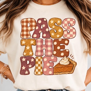 Puede incluir: Una sudadera de cuello redondo color crema con el texto PASS THE PIE en letras grandes con varios patrones. Las letras incluyen cuadros marrones y rosas, lunares y estrellas. Se incluye una porción de tarta de calabaza con nata montada.