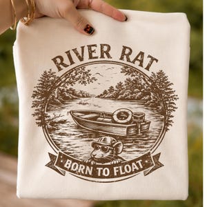 May include: Cream-coloured tote bag with a brown graphic. The graphic depicts a boat on a lake, trees, and the words "RIVER RAT" and "BORN TO FLOAT". A cartoon rat wearing sunglasses is also included in the design.