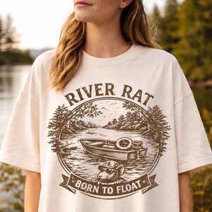 May include: Cream-colored t-shirt featuring a brown graphic with the words "RIVER RAT" above a boat scene. Below the boat is a mouse wearing sunglasses and the words "BORN TO FLOAT". The design is circular with trees and a river.