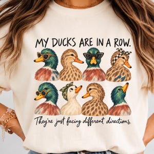 Könnte beinhalten: Ein cremefarbenes T-Shirt mit einer Grafik von acht Enten in zwei Reihen. Der Text lautet "MY DUCKS ARE IN A ROW." und "They're just facing different directions." Einige Enten tragen Accessoires wie eine Sonnenbrille mit Leopardenmuster, ein kariertes Stirnband oder ein grünes Bandana.
