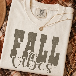 May include: A cream-colored short-sleeve t-shirt with a crew neckline and a Comfort Colors brand label at the inner neck. The front features the text FALL in large, distressed block letters and the word vibes in a cursive script, both in gray.