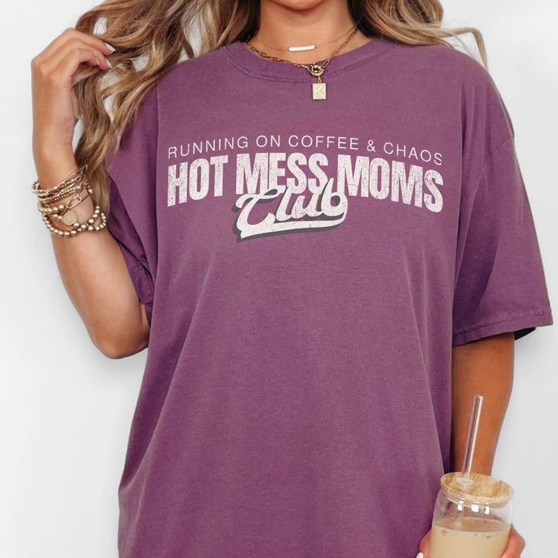 May include: A purple t-shirt with the text "RUNNING ON COFFEE & CHAOS HOT MESS MOMS Club" in white and green lettering. The shirt is a solid color and has short sleeves. The shirt is worn by a person.