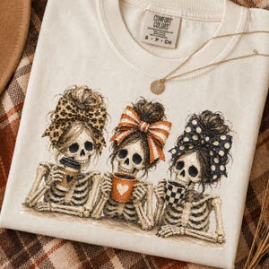 May include: Cream-colored t-shirt featuring a graphic of three skeleton figures with bows in their hair, holding coffee mugs. The bows have leopard print, orange stripes, and black polka dots. A gold necklace and the Comfort Colors tag are visible.