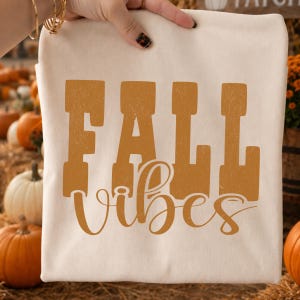 May include: A cream-colored t-shirt featuring the text FALL in large, bold, distressed mustard-yellow block letters, with the word vibes written underneath in a smaller, flowing, cursive mustard-yellow script font.