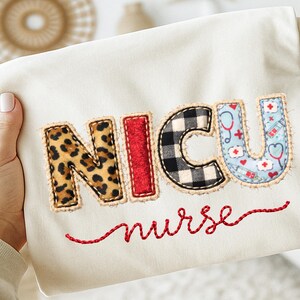 Puede incluir: Tela color crema con el texto bordado "NICU nurse". Las letras están aplicadas con varios patrones: estampado de leopardo, rojo, cuadros blancos y negros y un estampado azul con temática médica. La palabra "nurse" está cosida en rojo.