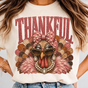 May include: A cream-colored t-shirt featuring a graphic of a turkey wearing leopard-print sunglasses, a pearl necklace, and a pink bow. The word THANKFUL is printed in large, textured, dark red letters above the turkey illustration.