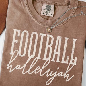 Può includere: Una t-shirt marrone a maniche corte con scollo rotondo ed etichetta del marchio Comfort Colors. La maglia presenta la scritta bianca FOOTBALL in un carattere serif grassetto e usurato, con la parola hallelujah in corsivo bianco sotto.