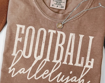 May include: A brown short-sleeve t-shirt with a crew neckline and a Comfort Colors brand label. The shirt features the white text FOOTBALL in a bold, distressed serif font, with the word hallelujah in a white cursive script font below it.