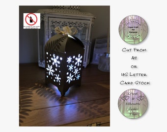 May include: A brown paper lantern with a snowflake design, lit with a white LED light. The lantern is cut from A4 or US Letter card stock. The image includes the text "THE SHADOW Paper Craft For Everyone" and "Cricut & Silhouette Cameo Files".