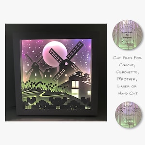 May include: A black shadow box with a white frame, featuring a paper cut design of a moon, stars, windmills, and houses. The design is illuminated from behind, creating a glowing effect. The text "Cut Files For Cricut, Silhouette, Brother, Laser or Hand Cut" is visible below the shadow box.