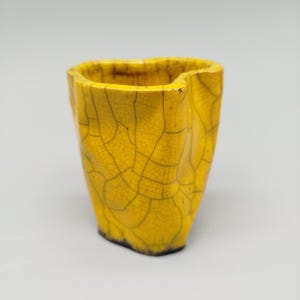 May include: A small, yellow ceramic vase with a unique, irregular shape. The vase has a cracked, textured surface with dark brown lines, and a dark brown base. The interior is also dark brown.