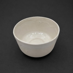 May include: A small, off-white ceramic bowl with a rounded base and a slightly flared rim. The interior surface features a glossy, reflective glaze, while the exterior has a matte, textured finish with subtle variations in the clay surface.
