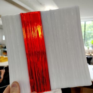 May include: A square, white glass plate with a textured surface and a vertical, vibrant red stripe down the centre. The plate is held by a hand, showcasing the contrast between the bright red and the white glass.