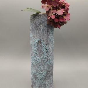 Op de afbeelding: Een hoge, cilindrische vaas met een textuur, blauwe en paarse glazuur. Een boeket roze hortensia's is in de vaas gerangschikt.