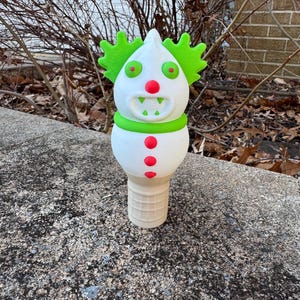 May include: A whimsical, 3D-printed clown figurine. The clown has a white head with green hair, red eyes and nose, and a green collar. The body features red buttons. The base is a waffle cone shape. The figurine is displayed outdoors.