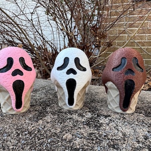 May include: Three Scream mask-shaped figurines in the form of ice cream cones. The ice cream flavors are pink, white, and brown, with black facial features. The cones are a light tan color. The figurines are displayed on a gray stone surface.