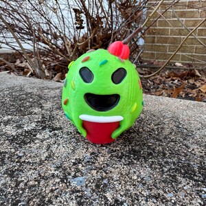 May include: A whimsical, green, spherical sculpture resembling a smiling face with black eyes and a wide open mouth. The sculpture has colorful sprinkles and a red cherry on top. It holds a red cup.