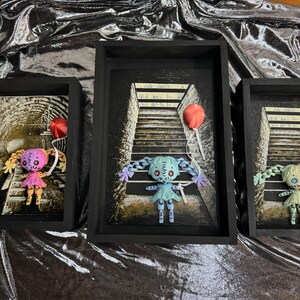 May include: Three black picture frames with white mats, each containing a doll with a balloon. The dolls are made of fabric and have stitched details. The dolls are posed on a staircase with a painted background. The balloons are red and are tied to the dolls' hands.