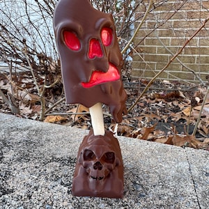 May include: A brown, skull-shaped sculpture with red accents, mounted on a wooden stick and a skull-detailed base. The sculpture has a melted appearance, with visible eye and mouth cavities. The base features a detailed skull design.