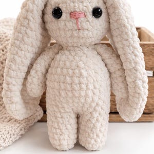 Peut inclure: Un lapin en peluche crocheté de couleur crème avec de longues oreilles tombantes, deux yeux ronds en plastique noir et un petit nez brodé en rose. Le jouet est fabriqué à partir d'un fil épais et texturé à l'aspect doux et moelleux.